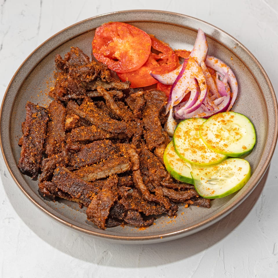 Spicy beef suya strips with sliced tomatoes, red onions, and cucumbers on a gray plate.