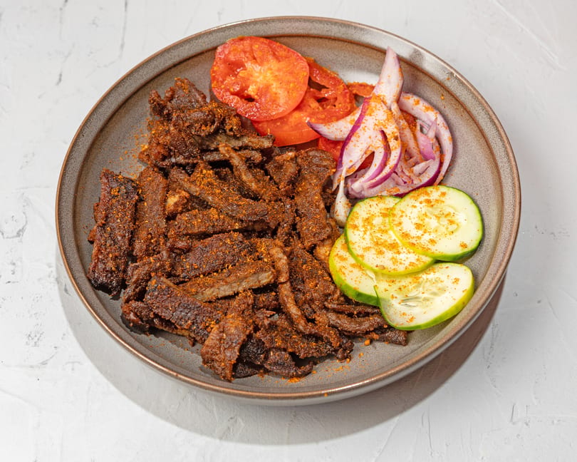 Spicy beef suya strips with sliced tomatoes, red onions, and cucumbers on a gray plate.