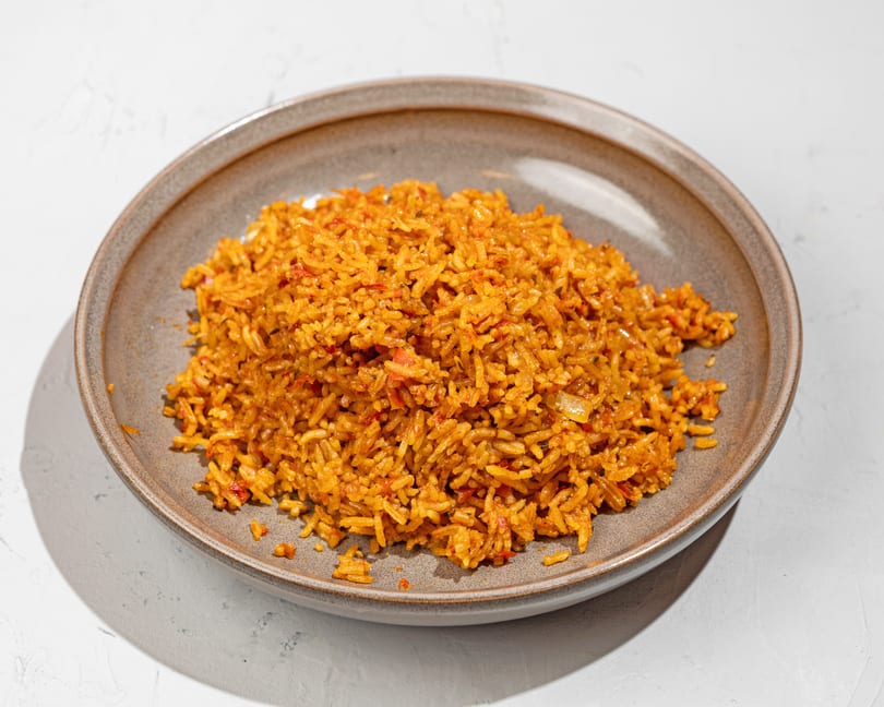 A large serving of vibrant orange jollof rice with red peppers in a rustic gray bowl.
