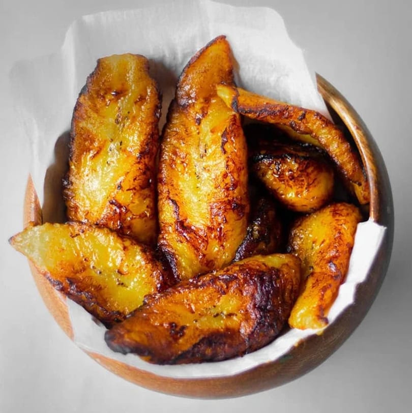 Golden brown fried plantains, some with darker caramelized edges, in a wooden bowl lined with white paper.